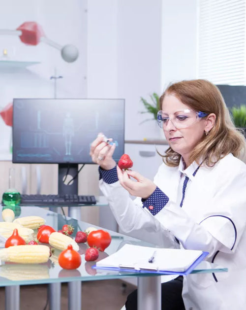 Scientist examining a red fruit in a laboratory setting with computer and vegetables on a table.